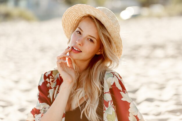 Fashion portrait of gorgeous blond woman with natural make up resting on sunny beach. Wearing straw hat. Holidays and vacation mood.