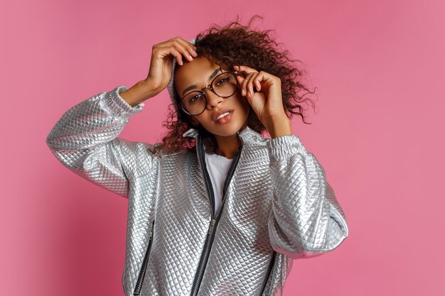 Fashion portrait of  mix race woman with brown skin and curly African hairstyle on vivid pink background. Wearing silver winter  jacket and grey hat.