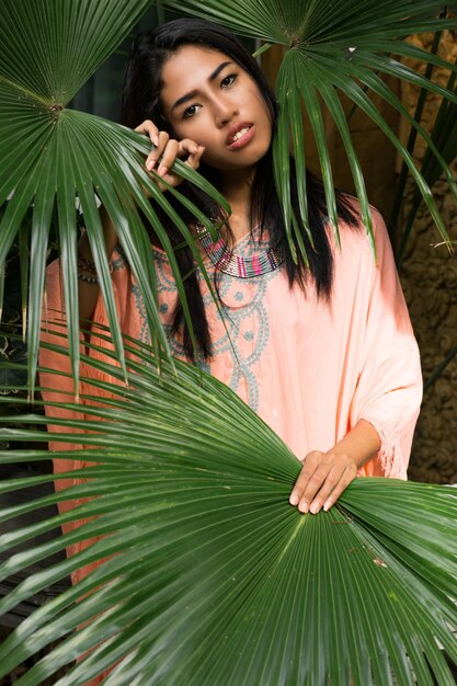 Fashion portrait of stunning asian woman posing in tropical garden. Wearing boho dress and stylish accessories.