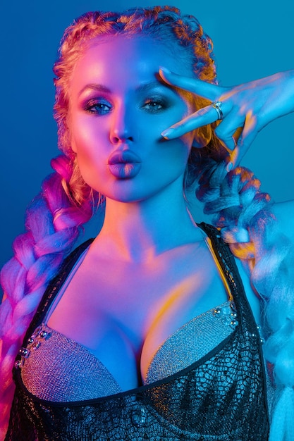 Fashion portrait of stunning sexy young blonde woman with trendy braids and sparkling bra under the net top She is holding peace sign by the eye making pouting lips in bright blue and red light