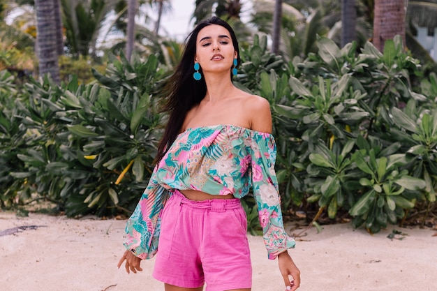 Free photo fashion portrait of stylish woman in colorful print long sleeve top and pink shorts on beach, tropical background.