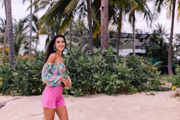 Free photo fashion portrait of stylish woman in colorful print long sleeve top and pink shorts on beach, tropical background.