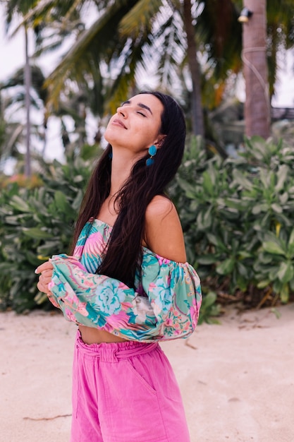 Free photo fashion portrait of stylish woman in colorful print long sleeve top and pink shorts on beach, tropical background.