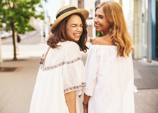 Fashion portrait of two young stylish hippie brunette and blond women models without makeup in summer sunny day in white hipster clothes posing . Turn around