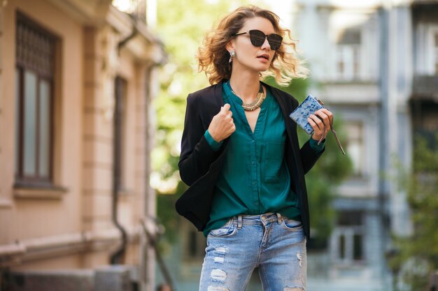 Fashion portrait of young elegant woman walking in street in black jacket, green blouse, stylish accessories, holding little purse, wearing sunglasses, summer street fashion style