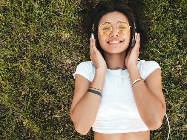 Free photo fashion portrait of young stylish hipster woman lying on the grass in the park .girl weares trendy outfit.smiling model enjoy her weekends. female listening to music via headphones.top view