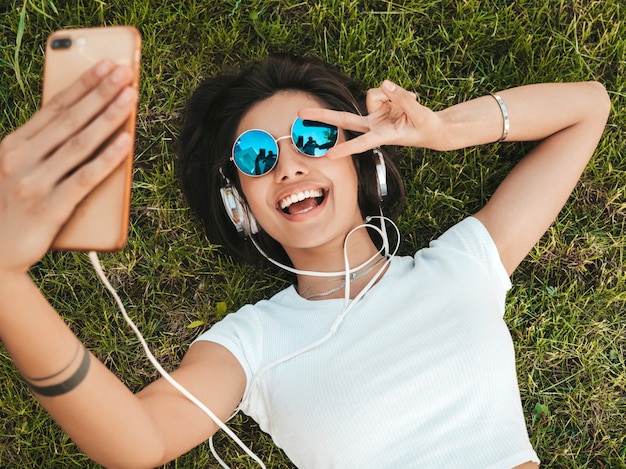 Free photo fashion portrait of young stylish hipster woman lying on the grass in the park .girl weares trendy outfit.smiling model making selfie. female listening to music via headphones.top view