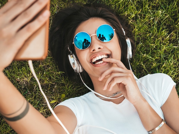 Free photo fashion portrait of young stylish hipster woman lying on the grass in the park .girl weares trendy outfit.smiling model making selfie. female listening to music via headphones.top view