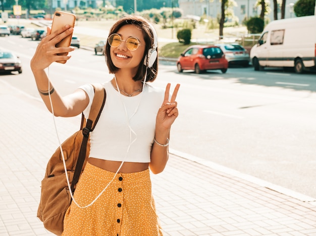 Fashion portrait of young stylish hipster woman walking in the street.Girl making selfie.Smiling model enjoy her weekends with backpack. Female listening to music via headphones