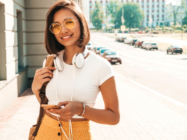 Fashion portrait of young stylish hipster woman walking in the street.Girl wearing cute trendy outfit.Smiling model enjoy her weekends, travel with backpack. Female listening to music via headphones