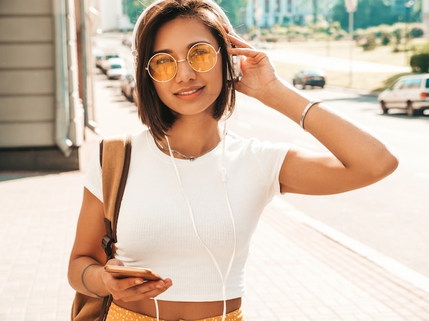 Fashion portrait of young stylish hipster woman walking in the street.Girl wearing cute trendy outfit.Smiling model enjoy her weekends, travel with backpack. Female listening to music via headphones