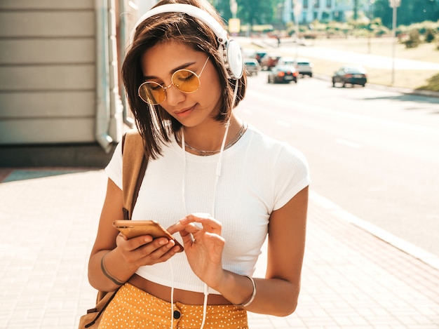 Fashion portrait of young stylish hipster woman walking in the street.Girl wearing cute trendy outfit.Smiling model enjoy her weekends, travel with backpack. Female listening to music via headphones