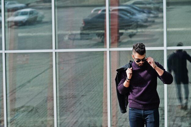 Fashion rich beard Arab man wear on turtle neck and sunglasses posed against large windows of modern building with jacket on hand Stylish succesful and fashionable arabian model guy