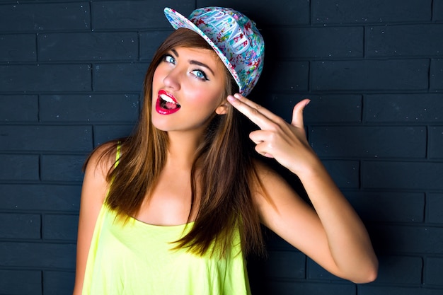 Free photo fashion sexy close up portrait of cheeky woman,imitating gun, put hand to her head, sexy full lips, open her mouth , swag bright hat, urban style, brick wall.