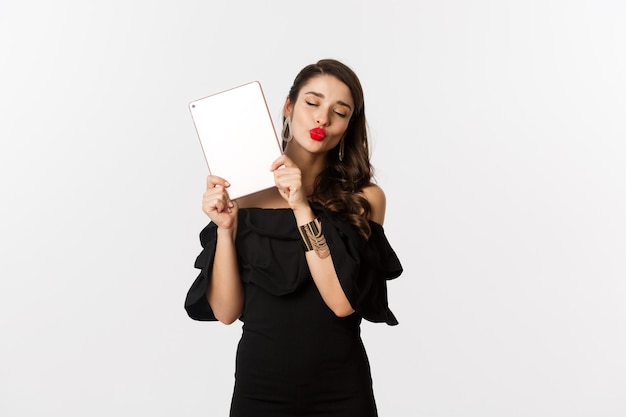 Fashion and shopping concept. Beautiful woman with red lips, wearing black dress, showing digital tablet and making kissing face, white background