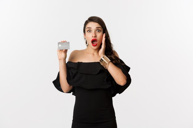 Fashion and shopping concept. Excited elegant woman with red lips, wearing black dress, showing credit card and looking amazed, standing over white background.