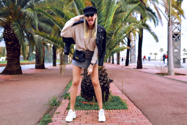 Free photo fashion street image of model with long legs wearing glamour stylish outfit, spring time, travel mood, palm trees around, sneakers, cap, leather jacket, mini skirt, sweater, accessories and jewelry.