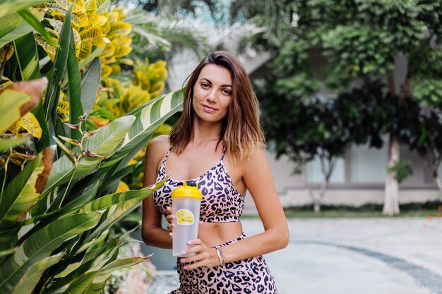 Fashion stylish young caucasian fit sporty woman in leopard cami top and biker shorts outside holds protein shaker