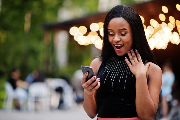 Fashionable african american woman in peach pants and black blouse look at mobile phone