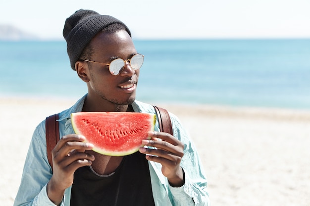 fashionable Afro-American man in black hat and sunglasses feeling happy and carefree holding big slice of rich red-ripe watermelon while spending summer day outside on urban beach