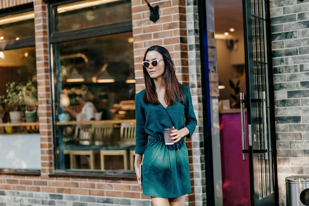 Free Photo fashionable attractive girl in summer green dress and sunglasses is walking in city park with coffee to go and spending weekend outdoor