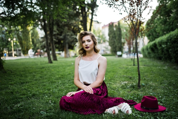 Free Photo fashionable and beautiful blonde model girl in stylish red velvet velour skirt white blouse and hat sitting on green grass at park