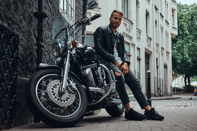 Free Photo fashionable biker dressed in a black leather jacket and jeans sitting on his retro motorcycle on old europe street.