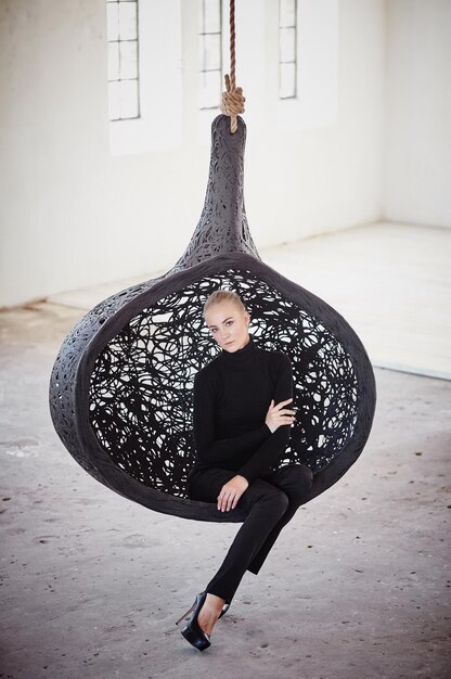 Fashionable blond female sits on a round chair in a big empty room.