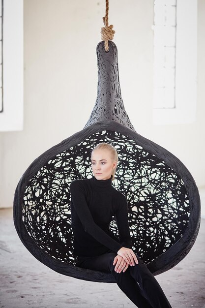 Fashionable blond female sits on a round chair in a big empty room.