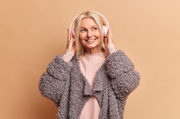 Free photo fashionable blonde european woman with pleasant smile wears stereo headphones enjoys favorite music has dreamy expression wears fur coat isolated over brown wall