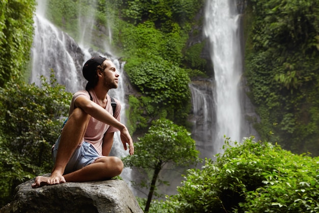 Free Photo fashionable caucasian hiker dressed casually sitting barefooted on big stone and relaxing during hard and long journey in rainforest. bearded male in snapback contemplating beautiful nature around him