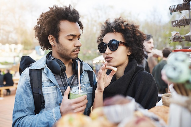 Free photo fashionable cute darkskinned couple in relationship with afro hairstyles standing near food counter in park picking something to eat and discussing group that will play today