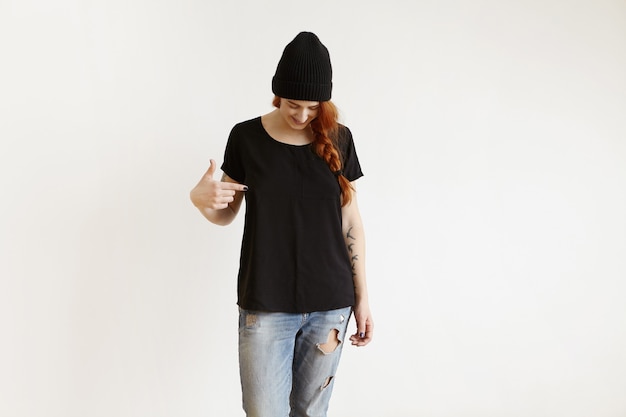 Free photo fashionable girl with braid posing indoors in black hat and ragged jeans, pointing her finger at t-shirt