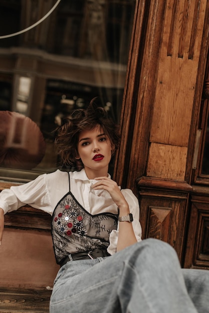 Fashionable lady in jeans with black lace and jeans poses on background of wooden door Curly girl with bright red lips looks into camera at street