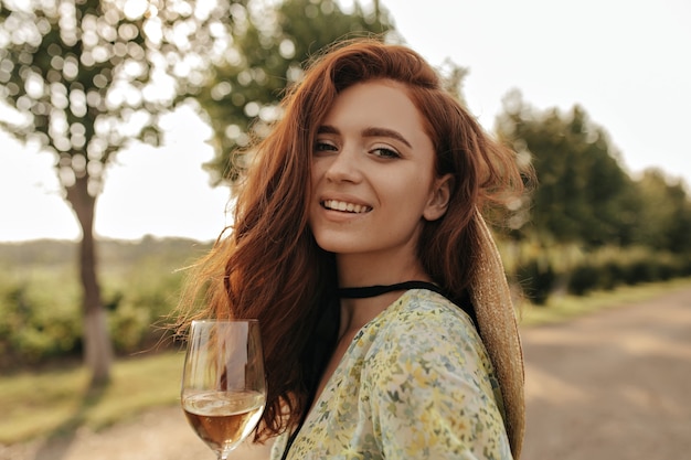 Fashionable lady with ginger wavy hairstyle and black bandage on her neck in modern green dress smiling and holding glass with drink outdoor