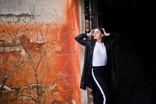 Fashionable long legs brunette model in long black cloak posed outdoor at winter day against old grunge wall