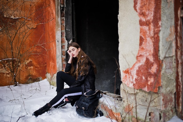 Fashionable long legs brunette model in long black cloak posed outdoor at winter day against old grunge wall