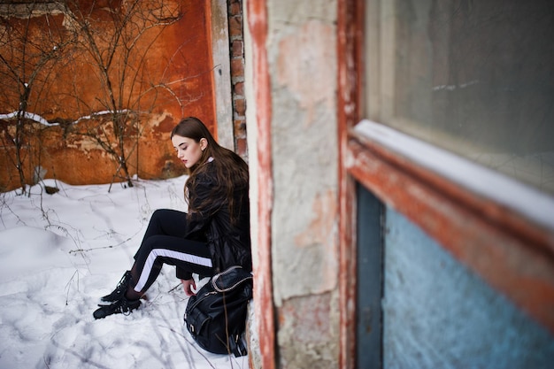 Fashionable long legs brunette model in long black cloak posed outdoor at winter day against old grunge wall