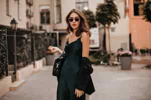 Free photo fashionable pale brunette in long green dress, black jacket and sunglasses, standing on street during daytime against wall of light city building