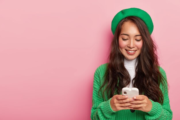 Free Photo fashionable pretty asian girl holds mobile phone, dressed in green clothes, surfes internet on modern cell phone, sends text message