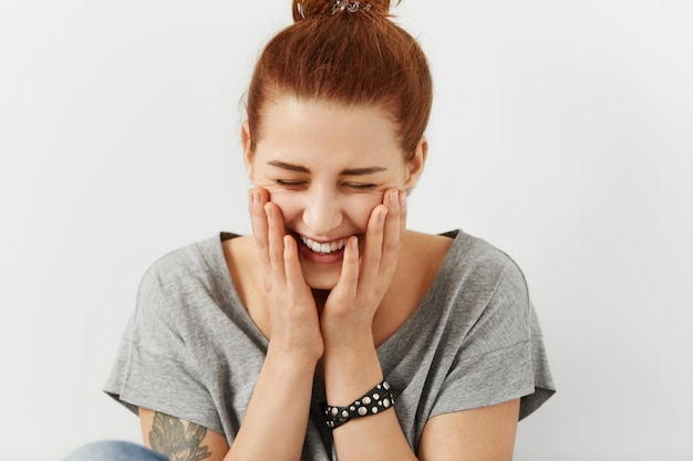 Fashionable pretty woman with hair knot and tattoo dressed in stylish grey t-shirt holding hands on cheeks and closing eyes