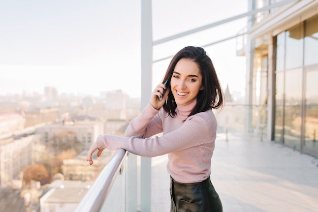 Free photo fashionable smiled young brunette woman on terrace in sunny day on big city view. talking on phone, laughing, having fun, success, relaxing time.