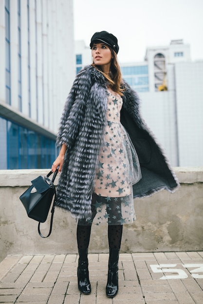 Fashionable smiling woman walking in city in warm fur coat, winter season, cold weather, wearing black cap, dress, boots, holding leather bag, street fashion trend