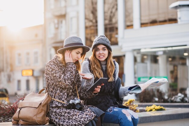 Fashionable smiling women sitting in the center of the city expressing brightful emotions on sunny day in city. Happy travelling together, trying to find the location on the map.