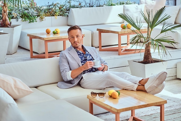 Free photo fashionable successful man with stylish hair dressed in modern elegant clothes holds cup of coffee while sitting on a sofa at outdoor cafe against the background of the city wharf.