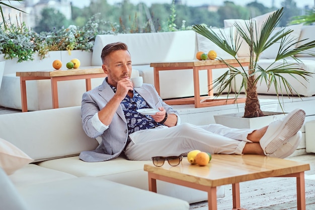 Free photo fashionable successful man with stylish hair dressed in modern elegant clothes holds cup of coffee while sitting on a sofa at outdoor cafe against the background of the city wharf.