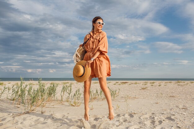 Fashionable summer image of beautiful brunette female in trendy linen dress , holding straw bag. Pretty slim girl enjoying weekends near ocean.Fell length.