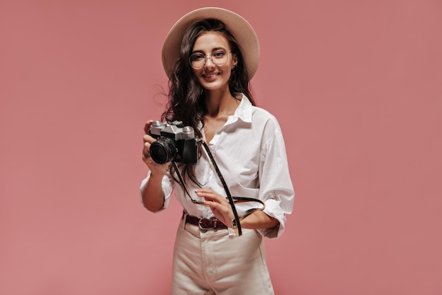 Free photo fashionable wavy haired lady in beige hat eyeglasses white shirt and modern pants smiling and posing with camera on pink background