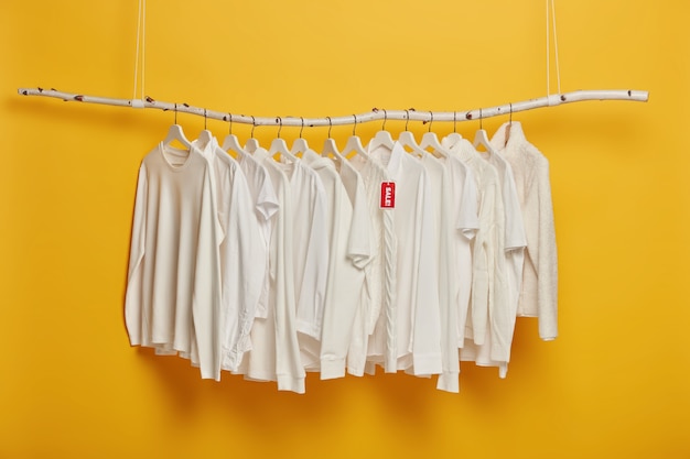 Fashionable white clothing on hangers with red tag inscriped sale, hanging on wooden rack against yellow background, copy space.