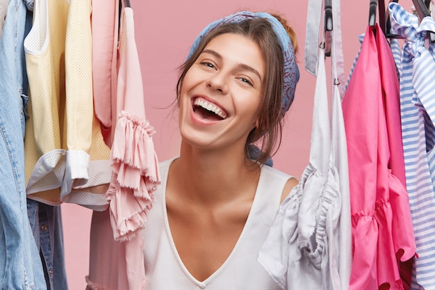 Fashionable woman choosing dress on date or party, feeling excited and happy. Cheerful woman having pleased look while packing bag before trip, standing in her wardrobe with racks full of clothes